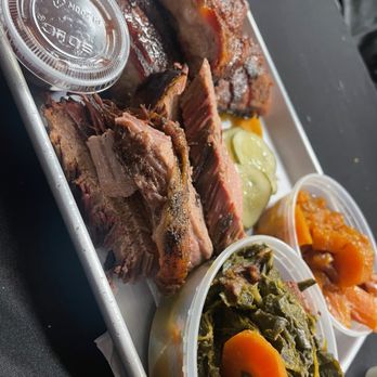 Backyard Barbeque - 2 Meat Combo: Pork Ribs, Beef Brisket, Yams, Collard Greens - A metal tray contains barbecue meats such as ribs and brisket, with sides of collard greens, pickles, and sauces, served on a dark table. - Barbeque Near Me - Freeport, New York