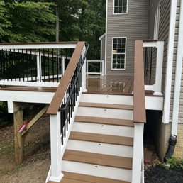 The image shows a newly constructed wooden deck with stairs leading up to it, attached to a residential house. The deck has a brown surface with white trim and black and brown railing. The stairs are brown with white sides, and the deck is elevated with support beams underneath. The house has beige siding and multiple windows, with a background of green trees and a partly cloudy sky.