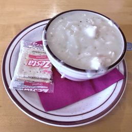 Green Chile Ground Sirloin - A bowl of green chile chicken stew containing chunks of chicken in a creamy, green sauce is shown on a white plate with brown trim. The plate also holds a saltine cracker and a pink paper napkin, all set on a wooden table surface. The stew appears thick and hearty, typical of comfort food served at a family-owned restaurant.