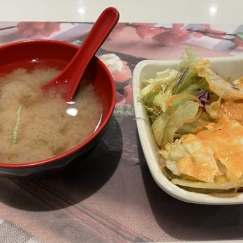 Bonsai 3 - Included with bento box - A bowl of miso soup with a red spoon and a small salad with dressing are placed on a table with a floral placemat. - Japanese Near Me - Fishkill, New York