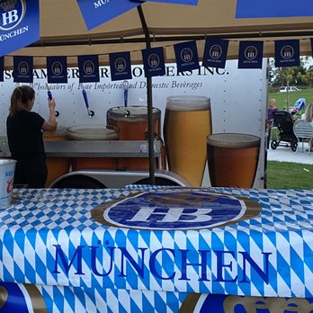 Hideaway Brew Garden - An outdoor beer stand with a blue and white checkered tablecloth displaying the Munich logo. The stand has several beer taps with large glasses of beer images on a backdrop. A woman is seen working behind the stand, and there are people and strollers in the background, indicating an outdoor event or festival setting. - Beer Gardens Near Me - Hoffman Estates, Illinois