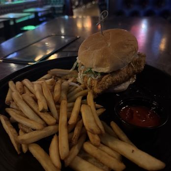 A.T. O'Keeffe's - Chicken sandwich - A fried chicken sandwich with lettuce and sauce in a bun is placed on a black plate alongside a serving of French fries and a small container of ketchup. The setting appears to be a dimly lit restaurant or bar with a wooden table and blurred background of patrons and neon lights. - Irish Pub Near Me - Boston, Massachusetts