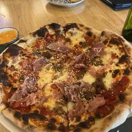 Carne pizza - A pizza with prosciutto, cheese, and vegetables sits on a white plate on a wooden table, with small side dishes and a black menu nearby.