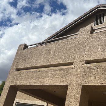 Mesa Roofing - A beige stucco building with a sloped roof, vent, and architectural openings, set against a partly cloudy sky. - Roofing Near Me