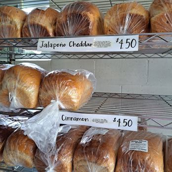 Pleasant View Bakery - Baked bread loaves are arranged on metal shelving units at Pleasant View Bakery. The loaves are wrapped in clear plastic and labeled with handwritten signs indicating flavors such as Jalapeno Cheddar, Cinnamon, and Honey Wheat, with prices ranging from $3.50 to $4.50. The background shows a plain wall and part of the bakery interior. - Bakeries Near Me - Marcellon, Wisconsin