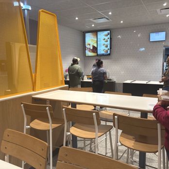 McDonald's - Fro t counter - The photo depicts the interior of a fast-food restaurant with a seating area featuring wooden chairs and tables. Yellow partition panels separate the seating from the ordering counters. Digital menu screens are mounted on the wall behind the counters, displaying food options. Several customers are standing in line or waiting, some holding drinks. The ceiling has recessed lighting, and the walls are tiled with a white, textured pattern. - Fast Food Near Me - Junction, Texas