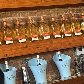 Cedar - Multiple bottles of Camarena liquor are lined up on a wooden shelf, with a brick wall as the background. Below, two champagne bottles are placed in light blue ice buckets labeled 'Lamarca Prosecco.' Additional bar tools and a small bottle are visible on the counter. The setting appears to be a bar or restaurant interior. - Breakfast & Brunch Near Me - Covington, Kentucky