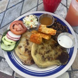 Pupusa Thursday! - A plate featuring Salvadoran pupusas with sides of fresh vegetables, pickled cabbage, and sauces, accompanied by drinks on a stone table.
