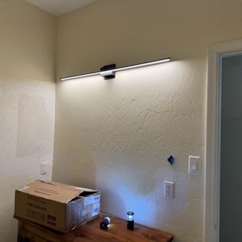 Wirez - New bathroom fixture - The image shows a room with a textured cream-colored wall illuminated by a sleek, horizontal wall-mounted light fixture. The fixture is turned on, casting light onto the wall. There is a wooden surface below with a cardboard box and two small flashlights. A switch and an electrical outlet are visible on the wall near the fixture, and part of a doorway is seen on the right side. - General Contractors Near Me