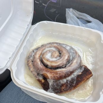 Pantry Restaurant - Nuff said... - A cinnamon roll with icing is shown in a white plastic takeout container inside a vehicle. The roll is spiral-shaped with icing on top, and the container is resting on a seat. The background includes part of the vehicle's dashboard, a plastic bag, and an electrical outlet with a purple charging cable. - Breakfast & Brunch Near Me - Santa Fe, New Mexico