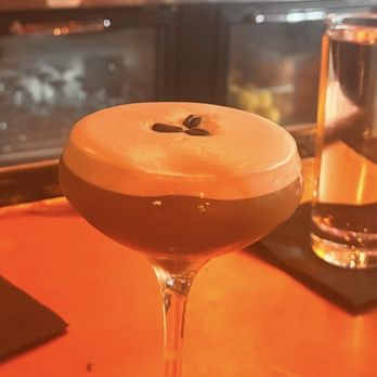 Sole's - Excellent white chocolate martini - A cocktail with a layered appearance, featuring a white foam top and a dark brown base, served in a coupe glass. The drink is garnished with two coffee beans on the foam. The setting appears to be a bar with a wooden surface, and a background of shelves with bottles and bar equipment, illuminated by warm lighting. - Bars Near Me - Keene, New Hampshire