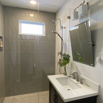 Belmore Glass & Mirror - A bathroom with a glass shower enclosure that has a handle and a small rectangular window above it. The shower area includes a multi-head shower system. Adjacent to the shower is a vanity with a white countertop, a built-in sink, and a dark cabinet below. A mirror is mounted on the wall above the vanity, flanked by two wall-mounted light fixtures. A plant in a glass vase and a soap dispenser are placed on the countertop. - Glass & Mirrors Near Me