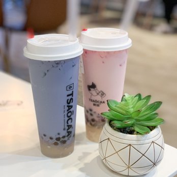 Tsaocaa - Taro & Jasmine - Two bubble tea drinks in plastic cups with lids and straws are positioned on a white table. One drink is purple with black tapioca pearls at the bottom, and the other is pink with similar tapioca pearls. A small, decorative potted succulent plant with green leaves is also on the table. The background is blurred, indicating an indoor setting, likely a bubble tea shop. - Bubble Tea Near Me - West Lafayette, Indiana