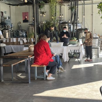 Sunday Bagels - Set up and operation - A group of people wearing masks sit and stand around a table in a brewery or taproom with brewing equipment in the background. - Bagels Near Me - Albuquerque, New Mexico