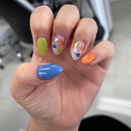 Close-up of a hand showing five manicured nails with different designs. The thumb nail is painted solid blue, the index nail is painted solid green, the middle and ring nails are clear with small, colorful heart-shaped stickers, and the pinky nail is painted solid orange. The background appears to be a salon or similar setting.