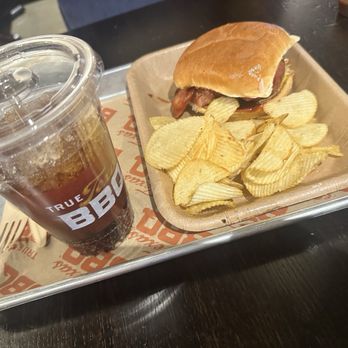 H-E-B - Sausage sandwich - A sausage sandwich with a bun and sausage link is placed on a tray alongside a serving of ridged potato chips and a plastic cup filled with a dark soda and ice. The tray is lined with branded paper from H-E-B, and the setting appears to be inside a grocery store or casual dining area in McKinney, Texas. - Grocery Near Me - Mckinney, Texas