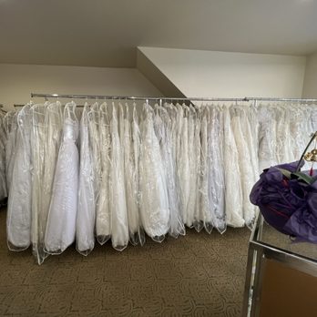 Fancy Wedding Center - 5/28/22 - Wedding Dress Shopping - A row of wedding gowns is hung on a metal clothing rack inside a bridal shop. The dresses are mostly white or ivory, some covered in clear plastic garment bags. The shop has a beige carpeted floor and a plain ceiling with recessed lighting. A small table or counter with a purple cloth and a decorative orchid is visible on the right side of the image. - Bridal Near Me - Brooklyn, New York
