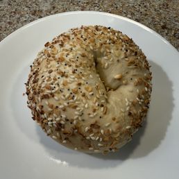 Everything Bagel - A round bagel with a hole in the center, spread with cream cheese and topped with sesame seeds, chopped nuts, and small black seeds, resting on a white plate. The plate is on a speckled granite countertop, and the image captures the bagel from a top-down perspective.