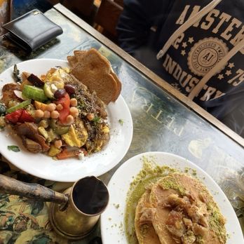 Brookline Lunch - The image shows two plates of Middle Eastern food on a table at Brookline Lunch in Cambridge, MA. One plate contains a vegetable and bean dish with bread, while the other features a sweet dessert topped with nuts and green powder. A small container of spice or sauce is also on the table. Part of a person wearing a dark hoodie with white text is visible, along with a wallet and a reflective table surface. The setting appears casual and cozy. - Diners Near Me - Cambridge, Massachusetts