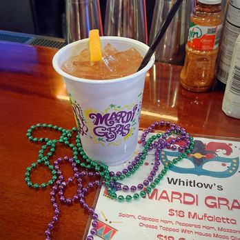 Whitlows - N'awlins Hurricane - A cold drink with ice cubes and a lemon wedge in a white cup decorated with Mardi Gras graphics, sitting on a bar counter. The counter has Mardi Gras beads, a menu, and various bar items in the background, including glasses and bottles. - Bars Near Me - Washington, DC, District of Columbia