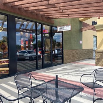 Boba Tealight - The image shows an outdoor seating area with metal chairs and a square table, situated on a concrete sidewalk in front of a cafe. The cafe has large glass windows displaying advertisements for drinks and snacks, with a neon 'OPEN' sign visible. A wooden pergola provides partial shade, and the building exterior includes green and beige walls with stone accents. Reflections of parked cars and trees are visible in the windows. - Bubble Tea Near Me - Gilbert, Arizona