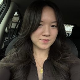 A woman with long, dark, wavy hair is seated inside a vehicle, taking a selfie. She has a neutral expression and is wearing a dark zip-up top and a gold necklace with a pendant. The background shows the interior of the car, including the headrest and window, with some outdoor scenery visible through the window.