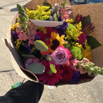 Niko's Flowers - A bouquet of various flowers including roses, sunflowers, and other colorful blooms is held by a person outdoors. The bouquet is wrapped in brown paper and is illuminated by sunlight, with a background of a sidewalk and parked cars. - Florists Near Me - Chicago, Illinois
