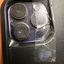 Scratches under the glass that weren't there before the replacement - The image shows the camera module of a smartphone with three lenses and a flash, covered by a clear plastic film that is peeling off.