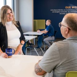 The Bank of Elk River - Main Street Office - A female banking specialist holding a branded The Bank of Elk River coffee mug while assisting a male client in a bright, ope...