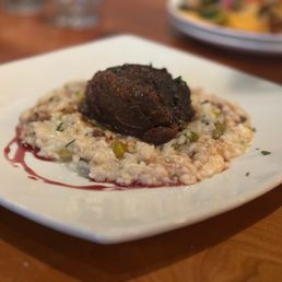 Beef Short Ribs - A plated dish displays a cooked beef short rib resting on a bed of risotto with visible green herbs and small vegetable pieces. The dish is served on a white square plate, with a drizzle of sauce around the risotto. The background shows a wooden table and other plates, indicating a restaurant setting.