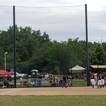 John Humphrey Complex - One field during tournament - The image shows a baseball game in progress on a field with players in uniforms gathered near the dugout area. Spectators are seated on benches and standing behind a fence, with some under a canopy. The background includes trees, parked cars, and a cloudy sky, indicating an outdoor setting during daytime. - Community Centers Near Me - Orland Park, Illinois