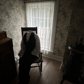 Door County Trolley - A mannequin without a head, dressed in a white shirt and black vest, is seated on a wooden chair near a window with lace curtains. - Wine Tours Near Me - Egg Harbor, Wisconsin