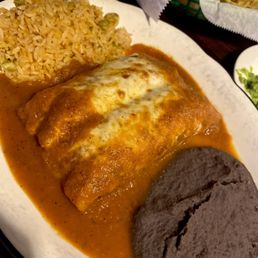 a plate of mexican food - A plate features an enchilada covered in sauce and melted cheese, served with rice and black beans, with chips visible in the background.