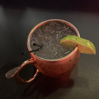 Bowlero Mesa - A copper mug filled with a dark beverage and ice cubes is shown on a dark surface. The mug has a handle and is garnished with a lime wedge on the rim. Two black straws are inserted into the drink, which appears to be a cocktail. The background is dark and plain. - Venues & Event Spaces Near Me - Mesa, Arizona