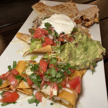 Taco Central - Central Sampler - A rectangular white plate displays Mexican cuisine, featuring tacos topped with chopped tomatoes, cilantro, and cheese, alongside a serving of guacamole, a dollop of sour cream, fresh salsa, and grilled tortillas. The dishes are arranged on a dark wooden table, highlighting the variety of ingredients and textures typical of Mexican food. - Beer Bar Near Me - Ansonia, Connecticut