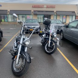 Arrived in the sun. Rode home in the rain. - The image shows two motorcycles parked side by side in a parking lot in front of Amherst Buffet, a large restaurant with a beige exterior and red sign. The motorcycles are wet from the rain, and the parking lot surface is also wet. Several cars are parked nearby, and the sky is overcast with gray clouds, indicating rainy weather.