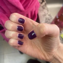 Nails - The image shows a close-up of a person's hand with nails painted in dark purple nail polish. Some nails have minor chips or uneven edges. The background includes a pink garment, possibly a sweater or jacket, and a blurred setting that appears to be indoors, with a surface and other objects partially visible.