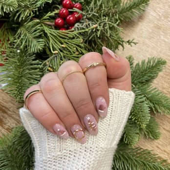 Trieu Beauty - A close-up image of a hand with decorated nails, featuring holiday-themed designs such as stars, moons, and Christmas trees, is held above a festive arrangement of evergreen branches and red berries. The hand is adorned with multiple rings and a white knit glove, set against a wooden surface background. The image emphasizes the manicure and jewelry in a seasonal setting. - Permanent Makeup Near Me - Albuquerque, New Mexico