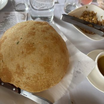 NiHao - Pancake - A large, round, fried bread item, likely a scallion bubble pancake, is placed on a white paper-lined tray. The pancake has a golden-brown, crispy exterior with some green flecks. It is accompanied by a serrated knife for cutting. The background shows a dining table with glasses of water, a bowl of fried rice, a cup of soup, and other tableware, indicating a restaurant setting. - Chinese Near Me - Baltimore, Maryland
