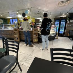new spot for lunch! - The image shows two customers standing at a cafe counter, with one wearing a black hoodie and the other in a beige sweater and cap. The cafe interior features a tiled floor, a wooden counter, and refrigerated beverage units. Seating includes black chairs with green cushions, and the ceiling has a patterned design with track lighting. The background includes menu boards and display cases.