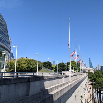 Chicago Bears - American flags are mounted on tall flagpoles along a concrete walkway in Chicago, adjacent to a modern glass building. The scene includes trees, a clear blue sky, and city skyscrapers in the distance, indicating an urban park or public space near a business district. - Professional Sports Teams Near Me