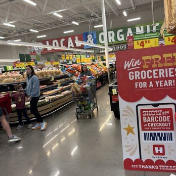 H-E-B - Produce - Customers are shopping in an H-E-B grocery store aisle, with a promotional sign offering a chance to win free groceries for a year. - Grocery Near Me - Austin, Texas