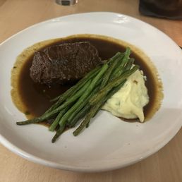 Short Ribs - The image shows a white, round plate with a serving of braised short rib in dark gravy, accompanied by green beans and a portion of mashed potatoes. The short rib is positioned on the left side of the plate, with the green beans laid across the middle and the mashed potatoes on the right. The dish is presented on a wooden table, with some utensils and a decorative cup visible in the background.
