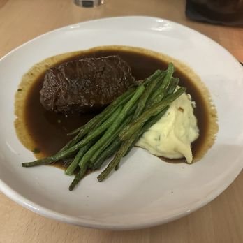 Caffeine's Midtown - Braised Short Rib - The image shows a white, round plate with a serving of braised short rib in dark gravy, accompanied by green beans and a portion of mashed potatoes. The short rib is positioned on the left side of the plate, with the green beans laid across the middle and the mashed potatoes on the right. The dish is presented on a wooden table, with some utensils and a decorative cup visible in the background. - American Near Me - West Springfield, Massachusetts