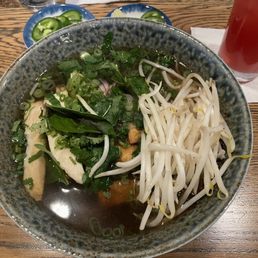 Vegan Pho - A ceramic bowl contains vegan pho with clear broth, topped with bean sprouts, chopped green onions, and fresh herbs such as basil. Sliced jalapeños are served on a small blue dish nearby. A glass of red-colored drink is placed on a white napkin to the side. The setting is on a wooden table, indicating a casual dining environment.