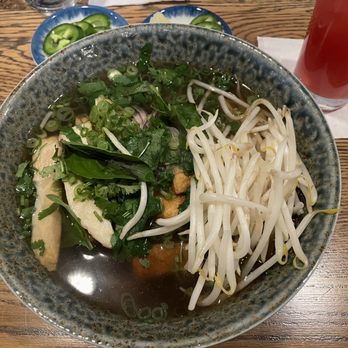 Pho Junkies - Vegan Pho - A ceramic bowl contains vegan pho with clear broth, topped with bean sprouts, chopped green onions, and fresh herbs such as basil. Sliced jalapeños are served on a small blue dish nearby. A glass of red-colored drink is placed on a white napkin to the side. The setting is on a wooden table, indicating a casual dining environment. - Food Trucks Near Me