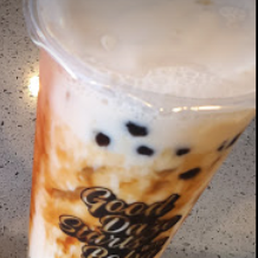 A clear plastic cup contains bubble tea with black tapioca pearls settled at the bottom. The drink appears to be a milk-based tea with a light, creamy color. The cup has a printed logo that reads 'Good Vibes Only' and is placed on a speckled countertop surface.