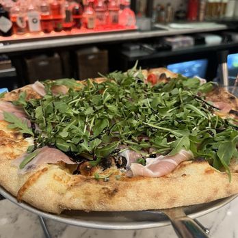 Bettola - Fig prosciutto Fig 'N' A Pizza - A pizza with a crust topped with fresh arugula, prosciutto slices, and drizzled with balsamic glaze, served on a metal stand. - Pizza Near Me - Cranston, Rhode Island