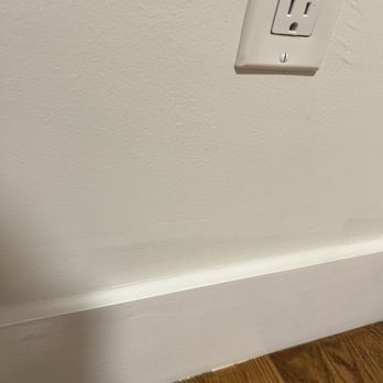 JW Cepp - Unfinished painting - The photo depicts a portion of an interior wall with a white electrical outlet cover mounted on it. Below the wall, there is a white baseboard running horizontally, with a hardwood floor beneath it. The wall has a textured surface, and the outlet cover is secured with a visible screw. The hardwood floor has a natural wood grain pattern and a polished finish. - Plumbing Near Me - Worcester, Massachusetts
