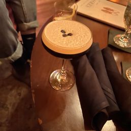 Espresso martini - A coffee cocktail served in a coupe glass, topped with a layer of foam and three coffee beans as garnish. The drink is on a dark wooden table, with a black napkin underneath. In the background, there is a glass with a lemon slice, a menu, and a decorative glass, indicating a bar or restaurant setting.